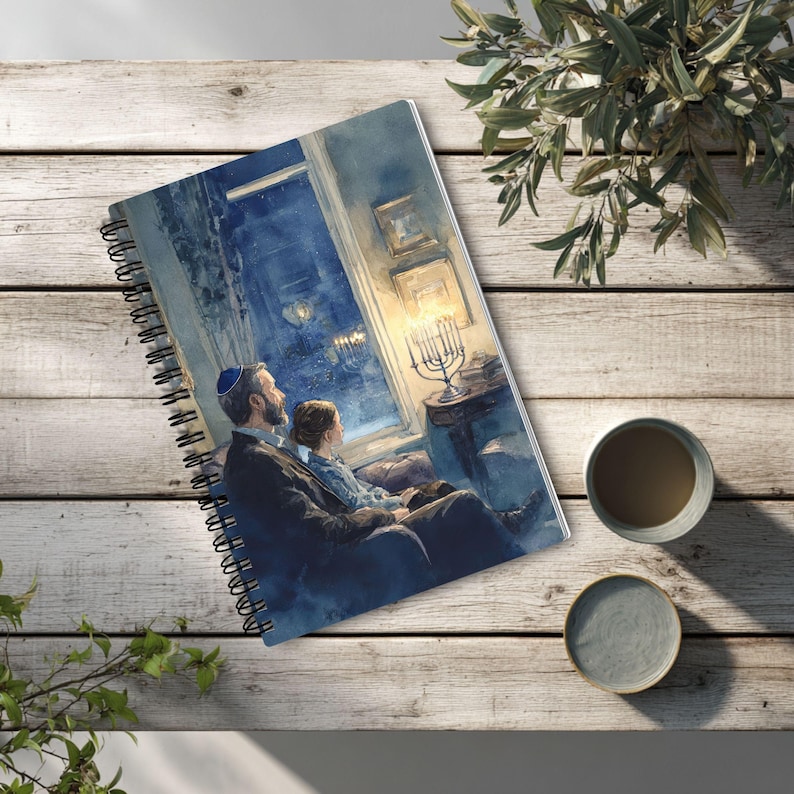 May include: A spiral-bound notebook featuring an illustration of a person wearing a blue kippah and a child sitting together by a window, looking at a lit menorah. The scene is rendered in shades of blue and warm yellow, capturing a quiet moment.