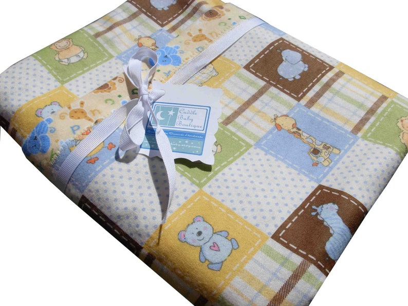 May include: A folded baby blanket featuring a patchwork pattern of squares in yellow, green, and blue with illustrations of teddy bears, giraffes, and elephants. A white ribbon is tied around the blanket with a tag labeled Cuddle Baby Boutique.