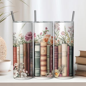 May include: Three stainless steel tumblers featuring a floral and book design. The tumblers depict a stack of vintage books with flowers growing around them. The tumblers are a light grey colour with a white background.