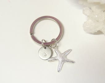 May include: A silver keyring featuring a starfish charm and a small, round charm with the letter "S". The keyring has a silver ring. The starfish charm is detailed and textured.