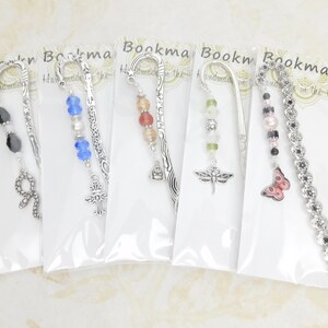 May include: A collection of five decorative bookmarks, each with a unique design. The bookmarks feature silver-toned metal accents and various charms, including beads, a cross, a dragonfly, a butterfly, and a snake. The bookmarks are packaged in clear plastic.