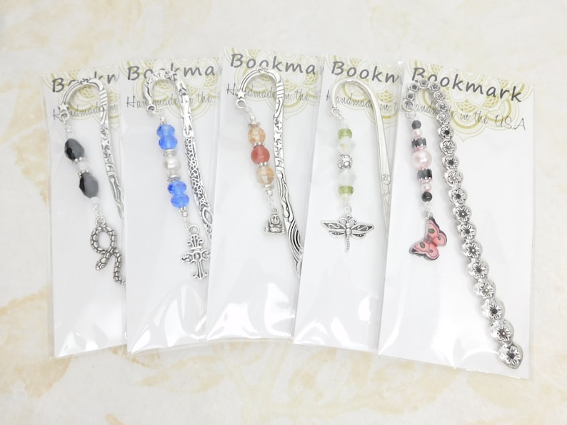 May include: A collection of five decorative bookmarks, each with a unique design. The bookmarks feature silver-toned metal accents and various charms, including beads, a cross, a dragonfly, a butterfly, and a snake. The bookmarks are packaged in clear plastic.