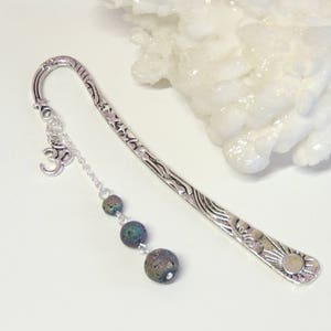 May include: Silver bookmark with a decorative design and a charm with the Om symbol. The bookmark has a chain with three dark green beads.