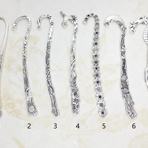 May include: A collection of seven silver-colored metal bookmarks, each with a unique design. The designs include floral patterns, sun and wave motifs, and animal figures such as a dolphin and a dragon. Each bookmark has a curved top with a small hole.