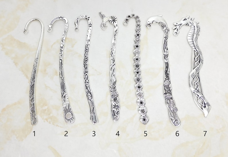 May include: A collection of seven silver-colored metal bookmarks, each with a unique design. The designs include floral patterns, sun and wave motifs, and animal figures such as a dolphin and a dragon. Each bookmark has a curved top with a small hole.