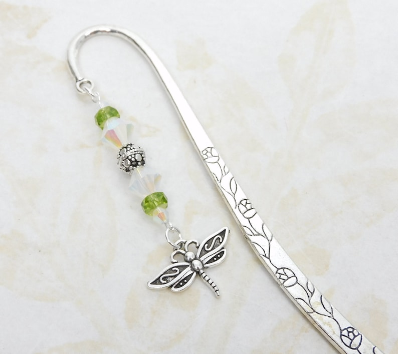 May include: Silver metal bookmark with a dragonfly charm and green and iridescent beads. The bookmark has a floral design along the edge. A decorative accessory for books.