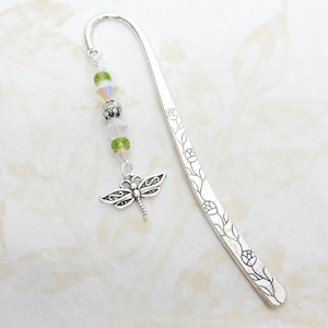 May include: A silver metal bookmark with a dragonfly charm and floral design. The bookmark features a curved top and a dangling charm with green and iridescent beads. The dragonfly charm is detailed with intricate wing patterns.