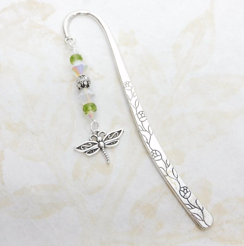 May include: A silver metal bookmark with a dragonfly charm and floral design. The bookmark features a curved top and a dangling charm with green and iridescent beads. The dragonfly charm is detailed with intricate wing patterns.