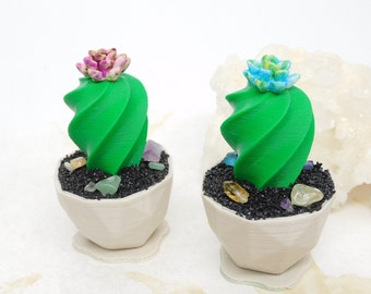 May include: Two decorative cactus sculptures. Each has a bright green, spiral-shaped body, topped with a colourful flower. They sit in off-white pots with black gravel and colourful crystals. The pots are on small, matching saucers.