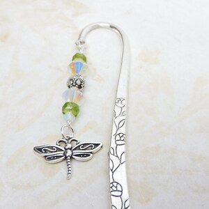 May include: A silver bookmark with a dragonfly charm and decorative floral etching. The charm is suspended from a strand of green and iridescent beads. The bookmark is made of metal and has a curved top.