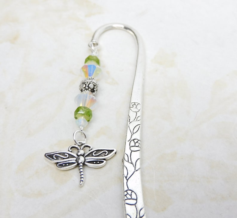 May include: A silver bookmark with a dragonfly charm and decorative floral etching. The charm is suspended from a strand of green and iridescent beads. The bookmark is made of metal and has a curved top.