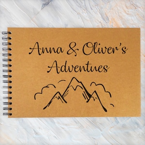Könnte beinhalten: Ein braunes Notizbuch mit schwarzer Spiralbindung. Der Umschlag zeigt den Text "Anna & Oliver's Adventures" und eine schwarze Strichzeichnung von Bergen.