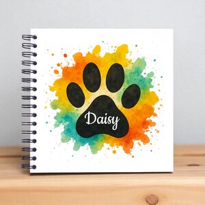 Könnte beinhalten: Ein spiralgebundenes Notizbuch mit einem schwarzen Pfotenabdruck-Design. Der Pfotenabdruck befindet sich vor einem farbenfrohen Aquarell-Spritzer aus Orange, Gelb und Türkis. Der Name "Daisy" ist in Weiß geschrieben.
