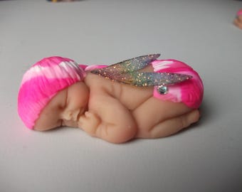 Könnte beinhalten: Miniaturfigur eines schlafenden Babys mit rosa und weißen Haaren. Das Baby hat einen hellen Hautton und ist mit glitzernden, schillernden Flügeln und einem kleinen Juwel verziert. Die Figur liegt auf der Seite.