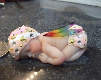 Könnte beinhalten: Eine kleine, schlafende Feenpuppe mit rosa und weißem Stoff, einem regenbogenfarbenen Stoffflügel und goldenen Glitzerakzenten.