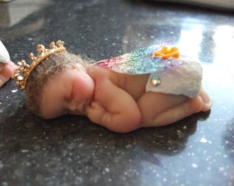 Könnte beinhalten: Eine schlafende Baby-Puppe, die ein weißes und regenbogenfarbenes Kleid mit einer goldenen Krone und einer gelben Blume trägt.