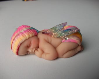 Könnte beinhalten: Eine schlafende Babyfigur mit einem regenbogenfarbenen Hut und glitzernden Flügeln. Das Baby ist nackt, mit einer rosa Windel und einem kleinen Juwel. Die Figur liegt auf der Seite, die Augen geschlossen.