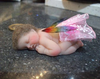 Könnte beinhalten: Eine kleine, schlafende Baby-Puppe mit einem rosa-weißen Stoffkleid und Regenbogenflügeln. Die Puppe ist aus Porzellan und hat ein realistisches Aussehen.
