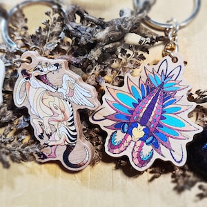 Peut inclure: Deux porte-clés en bois avec des illustrations colorées. Un porte-clés représente une créature de dessin animé avec des ailes et une queue rayée. L'autre porte-clés représente une créature colorée avec une longue queue plumeuse.
