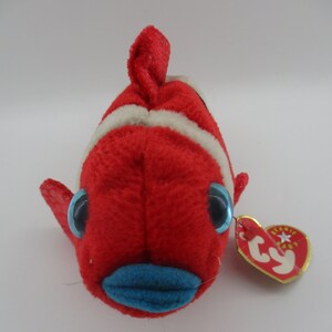 May include: Red and white striped beanie baby fish with blue eyes and a blue mouth. The fish has a white tag that says "Ty" with a star.