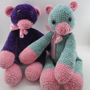 May include: Two handmade crochet teddy bears. One is purple with pink accents and a pink bow. The other is teal with pink accents and a pink bow. Both have black eyes and pink noses. The bears are sitting upright.