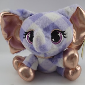 Puede incluir: Elefante de peluche morado y blanco con orejas y patas doradas. El elefante tiene ojos grandes y brillantes con párpados y pestañas morados.