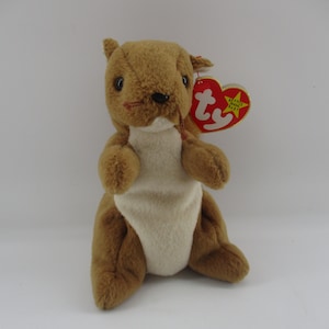 May include: A plush squirrel toy with brown fur and a cream-colored belly. It has small, black eyes, a red stitched mouth, and a red heart-shaped tag with yellow text. The toy is designed to sit upright.