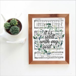 Può includere: Una stampa incorniciata con una ghirlanda floreale ad acquerello che circonda il testo "it is well with my soul". La stampa è su uno sfondo di spartito musicale vintage.