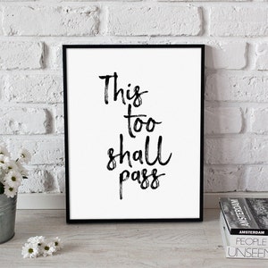 Puede incluir: Una impresión enmarcada con la frase "This too shall pass" en letras negras con pincel. La obra de arte se muestra sobre un fondo blanco y enmarcada en un marco negro. La impresión está apoyada contra una pared de ladrillos blancos.