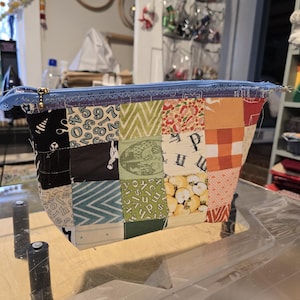 May include: A patchwork fabric zippered pouch with a blue zipper and gold-colored pull. The pouch is made of various fabric squares in black, white, green, orange, and floral patterns. The pouch is sitting on a wooden surface.