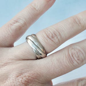 Może przedstawiać: Srebrny pierścionek z trzema splecionymi obręczami. Pierścionek jest noszony na palcu.