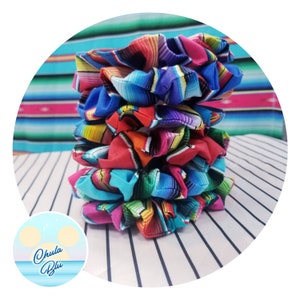 May include: A stack of colorful scrunchies with a vibrant striped pattern in shades of red, blue, green, and orange. The scrunchies are made of fabric and are stacked on top of each other. The background features a striped pattern.