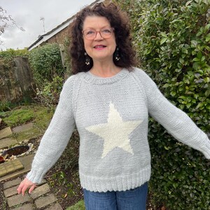 May include: A light gray long-sleeved knit sweater featuring a large, centered cream-colored star design on the front. The garment has a ribbed crew neckline, ribbed cuffs, and a ribbed hem, constructed with a soft, textured yarn material.