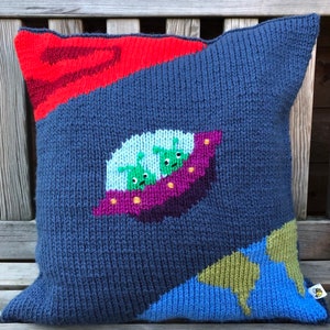 May include: A knitted pillow with a blue background and a purple spaceship with three green aliens inside. The spaceship is flying over a red and blue planet. The pillow is made with a textured knit.