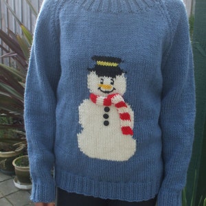 May include: A blue knitted jumper with a snowman motif. The snowman has a black top hat, a red and white striped scarf, and black button eyes. The jumper has a ribbed neckline, cuffs, and hem.