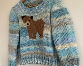 May include: A blue and white striped knitted sweater with a brown bear applique. The sweater has a crew neck and long sleeves.