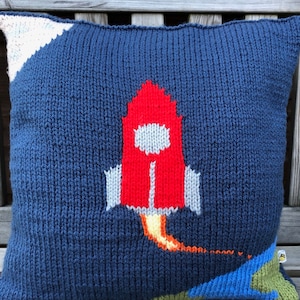 May include: A blue knitted pillow with a red, white, and yellow rocket ship design. The rocket ship is flying through the air with a trail of smoke behind it.