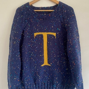 Peut inclure: Un pull en tricot bleu marine avec une lettre "T" jaune sur le devant. Le pull a un motif moucheté avec des points multicolores.