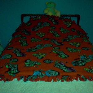 May include: A red fleece blanket with a repeating pattern of motorcycles in various colors. The blanket is on a bed with a white comforter and a wooden headboard. A teddy bear sits on top of the headboard.