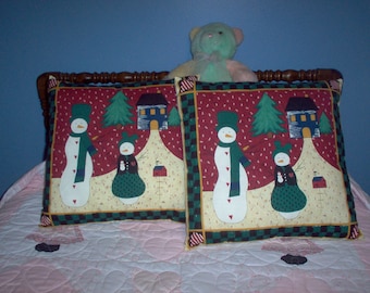 May include: Two decorative square pillows with a winter scene featuring snowmen, houses, and trees. The pillows have a red background with a green and black checkered border. A stuffed teddy bear sits behind the pillows.