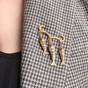 May include: A gold-toned brooch featuring three fish pendants suspended from a curved bar. The fish have detailed scales and fins. The brooch is pinned to a brown and white houndstooth patterned blazer.