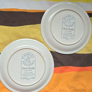 May include: Two off-white Hearthside stoneware plates. The plates have a blue logo that reads "Casual Elegance" and "Hearthside Stoneware". The plates are marked "Safe in your dishwasher & microwave oven" and "Chablis 571 Japan".
