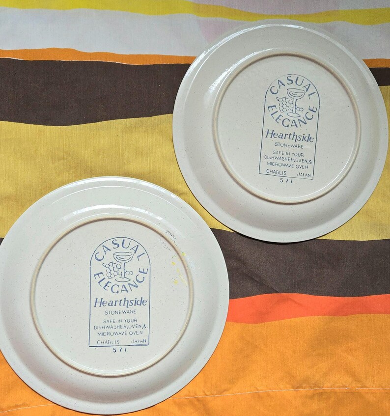May include: Two off-white Hearthside stoneware plates. The plates have a blue logo that reads "Casual Elegance" and "Hearthside Stoneware". The plates are marked "Safe in your dishwasher & microwave oven" and "Chablis 571 Japan".