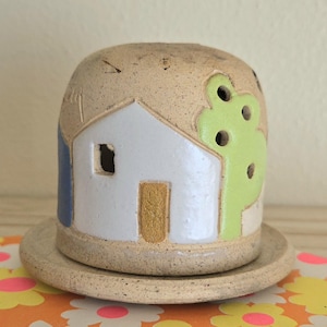 May include: A ceramic house-shaped candle holder with a matching saucer. The house is painted white with a yellow door, blue side, and a green tree. The candle holder has a textured, beige finish.