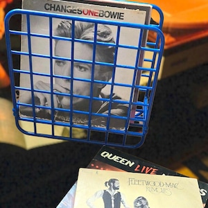 May include: A blue wire record holder filled with vinyl records. The top record features a black and white image of a man and the text "CHANGESONEBOWIE". Other records include "QUEEN LIVE" and "Fleetwood Mac Rumours".