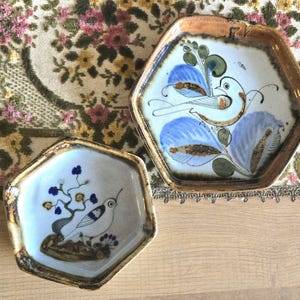 May include: Two hexagonal ceramic dishes with hand-painted bird and floral designs. The dishes feature a white base with blue, brown, and green accents. The rims have a textured, brown finish. One dish has a bird with blue leaves.