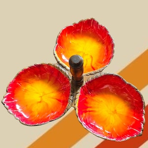 May include: A three-section serving dish featuring three leaf-shaped bowls with a vibrant red and yellow gradient finish. The bowls are connected at the center by a dark brown, cylindrical wooden handle with a textured, rope-like metal base.