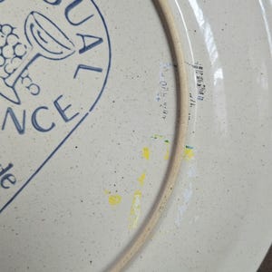 May include: A section of a white ceramic plate with a blue design and text. The design includes a wine glass and grapes. The plate has a speckled texture and a light brown rim. The text reads "PROVENCE" and "ide".