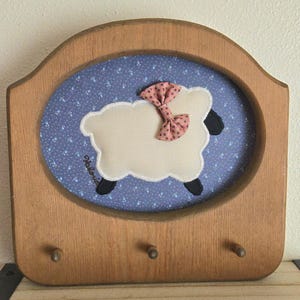 May include: A wooden wall-mounted hook rack featuring an oval inset with a blue fabric background patterned with small white flowers. A cream-colored fabric sheep appliqué with a pink polka-dot bow is centered, with the name Valerie embroidered.