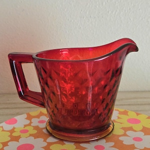 May include: A vibrant red glass creamer pitcher featuring a textured diamond pattern across its exterior surface. The vessel includes a sturdy angular handle on one side and a prominent pouring spout, showcasing a translucent finish throughout.